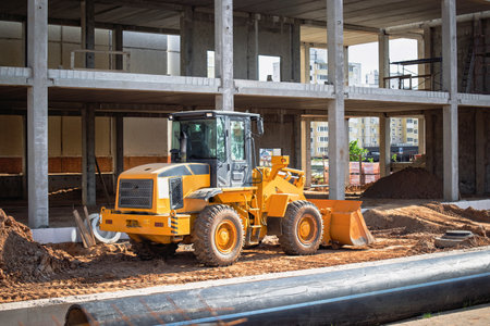 A yellow loader is actively engaged in moving earth at a construction site. The backdrop features an incomplete building structure under a clear sky.の写真素材
