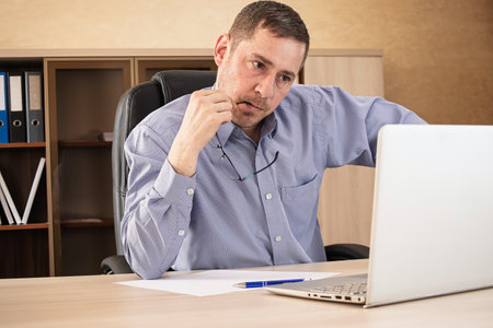 A man in a blue shirt sits at a desk, deep in thought while working on his laptop. Papers and a pen are nearby as he contemplates his tasks.の写真素材