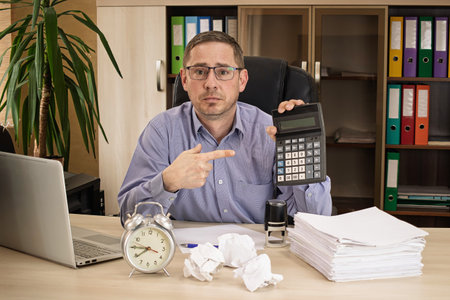 An office worker with glasses shows visible frustration as he points at a calculator while surrounded by crumpled paper and a ticking clock on his desk.の写真素材