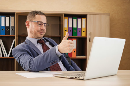 A man in a suit sits at a desk in a professional office, smiling and giving a thumbs up after completing a task on his laptop.の写真素材