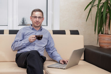In a comfortable living space, a man sits on a couch, sipping coffee from a cup while focused on his laptop. Green plants add warmth to the room.の写真素材