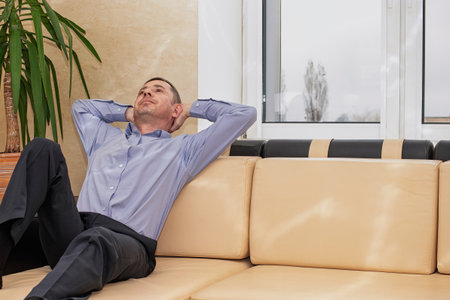 A man sits comfortably on a light-colored couch, leaning back with his hands behind his head, taking a moment to relax indoors.の写真素材