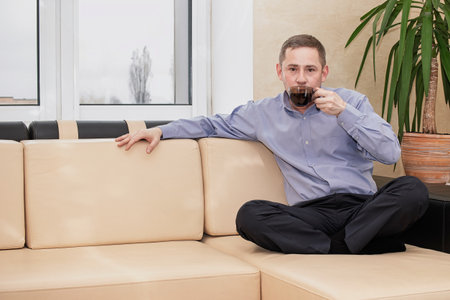 A man sips coffee while seated cross-legged on a beige couch in a cozy indoor setting. The atmosphere is relaxed with minimalistic decor.の写真素材
