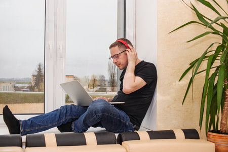A man is focused on his laptop, wearing headphones, and sitting comfortably by a window in a contemporary apartment on a cloudy day.の写真素材