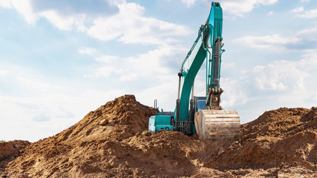 Excavator is operating on a construction site. It digs into a large pile of soil under a clear blue...