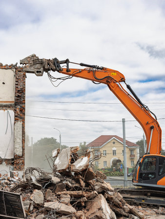 A strong excavator is actively dismantling a building as debris falls to the ground. Dust clouds rise, showing the ongoing demolition work in the urban setting.の写真素材