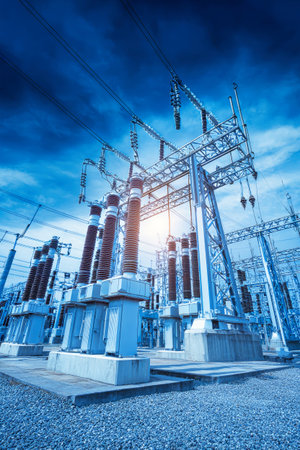 Electricity flows through a large substance in a rural area as the sun sets. Towering electricity poles and transformers are visible against the sky.の素材