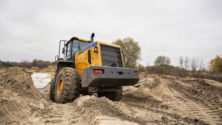 A large yellow tractor is digging in sandy soil at a construction site. The cloudy sky and autumn...