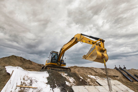 A yellow excavator lifts soil at a construction site surrounded by piles of dirt, with dark clouds...