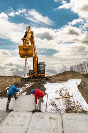Two construction workers are carefully laying concrete slabs while a large excavator operates in the...