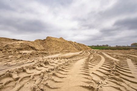 Deep tracks from heavy machinery cut through the sandy ground on a cloudy day. A large pile of sand rises in the background, hinting at ongoing construction.の写真素材