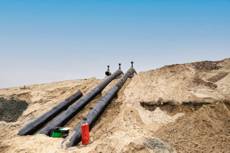 Large black pipes are placed on a sandy construction site. The clear sky and sandy landscape...
