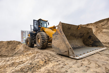 A large loader works at a construction site, moving soil with a bucket in front of a building in an...