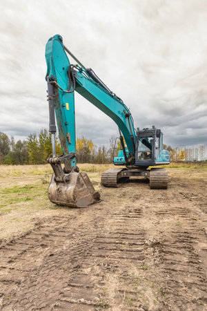 Excavator digs on a construction site with trees in the background under a cloudy sky during autumn,...