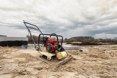 A compacting machine uses vibrations to compress sand on a construction site. Workers prepare the...