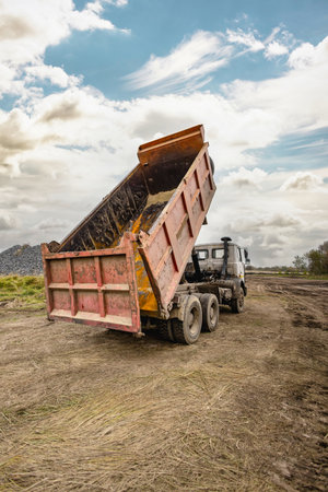 A dump truck is unloading a load of dirt at a construction site surrounded by a rural landscape. It is an overcast day with visible clouds.の写真素材
