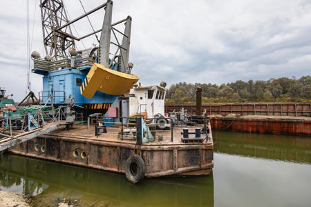 A large barge with construction equipment is docked at a waterway, surrounded by trees, under cloudy...