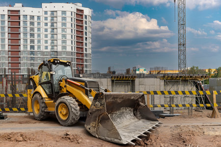 A backhoe loader sits at a construction site, preparing the ground next to a partially completed tall building as storm clouds gather overhead.の写真素材