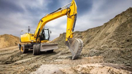 A large yellow excavator digs into the sandy ground at a construction site. Dark clouds loom...