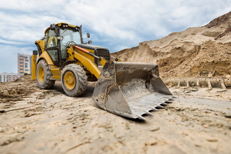 A yellow excavator moves through a construction site, skillfully digging and moving dirt against a backdrop of tall buildings and an overcast sky.の写真素材