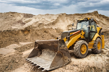 A yellow backhoe loader works on a construction site, moving dirt and debris while clouds gather...