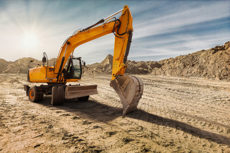 Heavy machinery digs into the dry soil of a construction site. The sun sets, casting warm hues over the landscape and highlighting the excavator's powerful arm.の写真素材