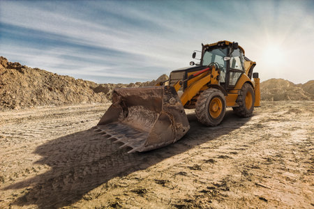 A yellow backhoe loader digs into the earth on a construction site as the sun sets, casting long shadows on the rugged ground.の写真素材