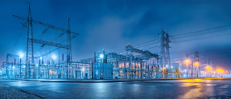 At night, an electrical substance stands illuminated with bright lights highlighting its structures and power lines against a calm urban background.の素材