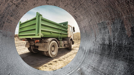 A construction site reveals a green truck parked on rough terrain, framed by a large pipe, with a blue sky stretching above and construction activities in motion.の写真素材