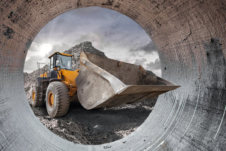 Excavator lifts dirt at a bustling construction site as clouds gather overhead, showcasing the hard work and equipment in action from inside a large pipe. Loader, excavatorの写真素材