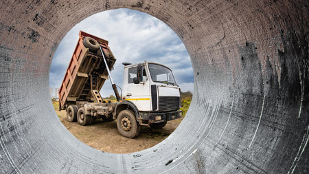 Construction equipment is busy unloading materials at a site, viewed through a large pipe structure under a cloudy sky, highlighting ongoing activities.の写真素材