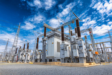 Large electrical substation filled with high voltage equipment and power lines, set against a backdrop of a clear blue sky and fluffy white clouds in daylight.の素材