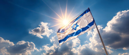 In a stunning display, the flag of Israel waves proudly against a bright blue sky filled with white clouds and sunlight illuminating the scene.の素材
