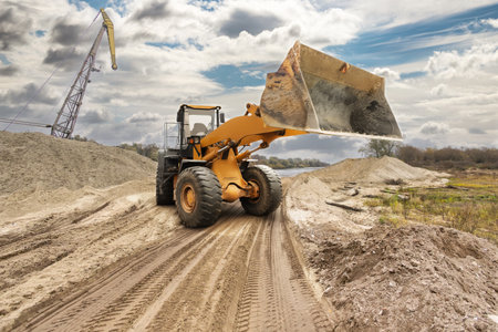 Loader lifts sand at a construction site beside a river. Clouds drift overhead as workers prepare the area for future projects.の写真素材