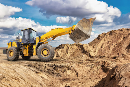 A large yellow loader works on a construction site, lifting and moving dirt alongside a sandy hill in bright daylight.の写真素材