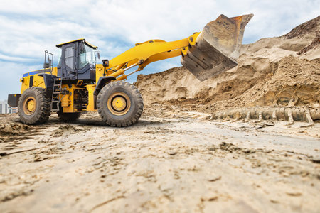 A large yellow A wheeled bulldozer or loader is moving sand at a construction site, working efficiently under a partly cloudy sky with a city backdrop.の写真素材