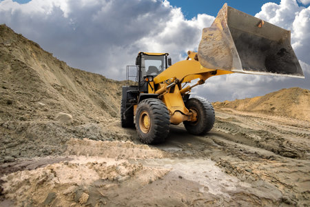 A large loader is lifting dirt at a construction site surrounded by mounds of soil under a dramatic sky in the afternoon light.の写真素材