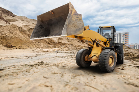 A loader is seen lifting a heap of dirt at a construction site in a city, surrounded by tall buildings and a cloudy sky during the day.の写真素材