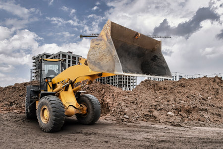 A large bulldozer loader lifts a pile of dirt on a construction site surrounded by buildings and cranes under a cloudy sky during the day.の写真素材