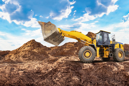 A yellow A wheeled bulldozer or loader lifts a large scoop of dirt at a construction site. The bright sky and piles of earth create a busy atmosphere in the daytime.の写真素材