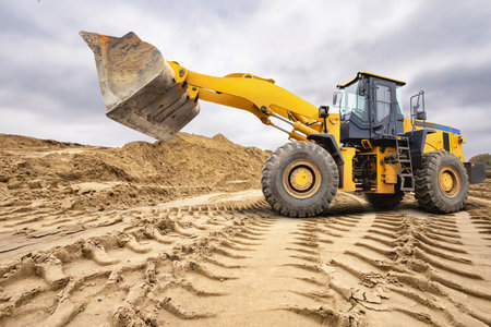 A large yellow loader excavator lifts a heavy load of soil at a construction site. Thick clouds cover the sky, indicating possible rain.の写真素材