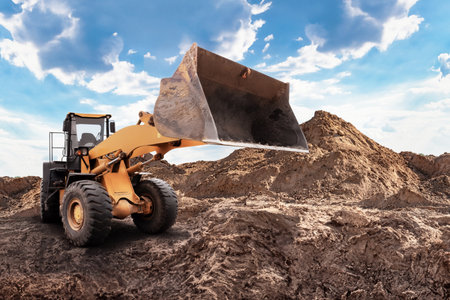 A loader lifts soil at a construction site surrounded by mounds of dirt under a bright blue sky with scattered clouds.の写真素材