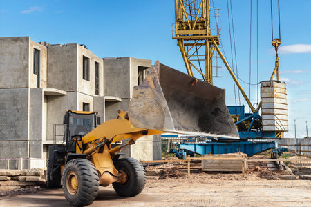 Heavy machinery is actively engaged on a construction site, with a loader moving dirt and a crane positioned to lift materials against a blue sky.の写真素材