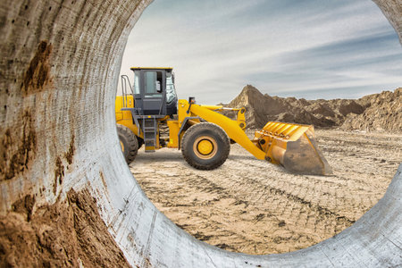 A large yellow loader moves dirt on a construction site. It is framed by a concrete pipe, showing an...