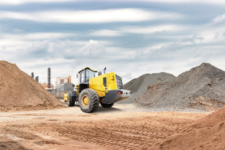 Construction equipment moves through a site filled with large mounds of dirt and gravel. Industrial...