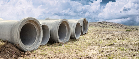 Concrete pipes are placed on dry land surrounded by grass and dirt, with a cloudy sky above. The...