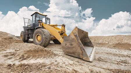 A loader works on a construction site, digging and moving soil as clouds fill the sky. The machinery...