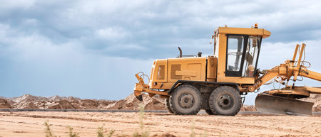 A yellow grader is actively leveling the sandy terrain at a construction site while dramatic clouds...