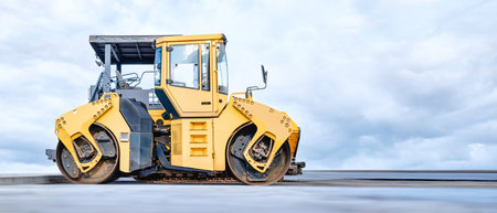 A bright yellow roller compactor works hard at a busy construction site. Dark clouds hang overhead,...