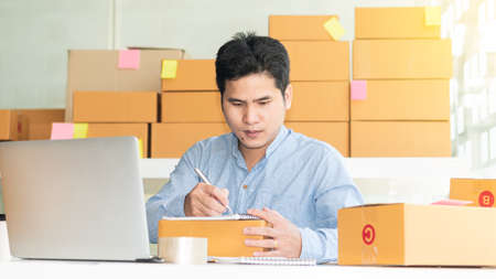 A young Asian businessman works at home to sell stuff online and takes note of the order for successful delivery in the online store.の写真素材